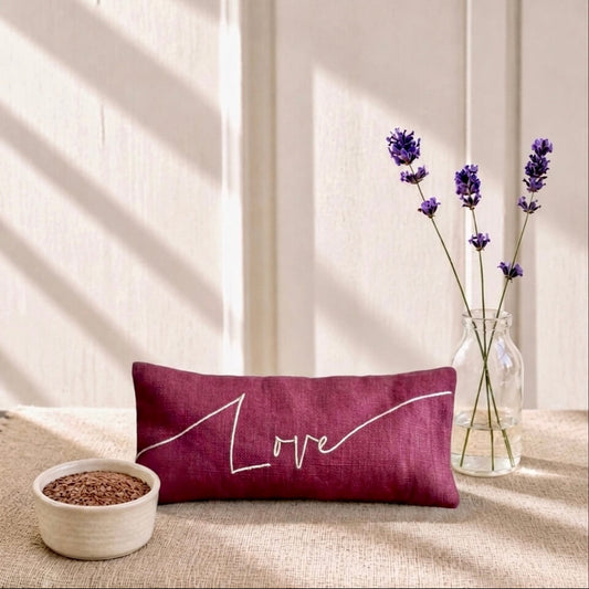 Red pillow with 'love' text on a textured surface with a vase of flowers and bowl in the background.