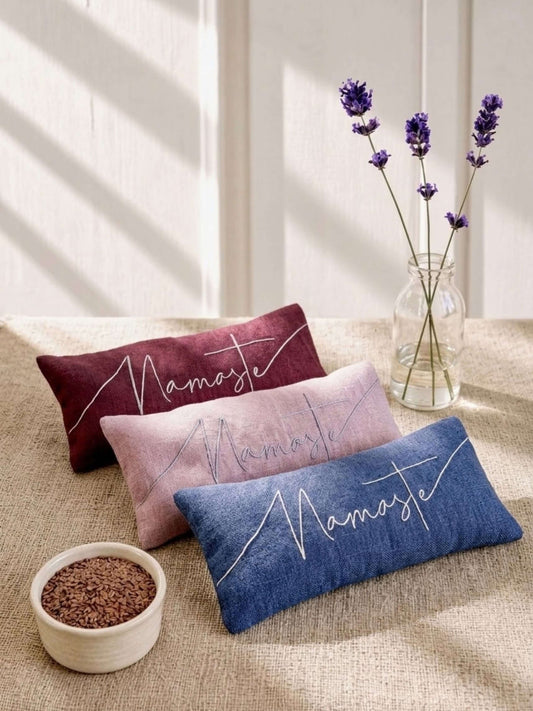Three rectangular decorative eye pillows in red, dusty pink, and blue linen, each embroidered with the word 'Namaste' in white script, arranged on a beige fabric surface beside a small bowl of brown beans and a glass vase with purple lavender stems.