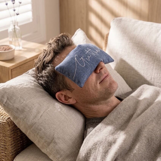 A man lying in a bed with pillows and a blanket, wearing a blue eye pillow with white script text that reads 'Gratitude'.