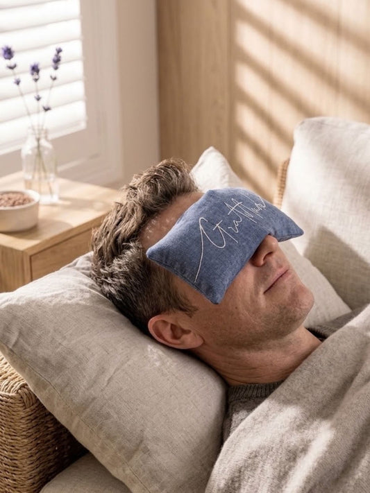 A man lying down under a blanket resting his head on a pillow, wearing a blue eye pillow embroidered with the word Breathe.