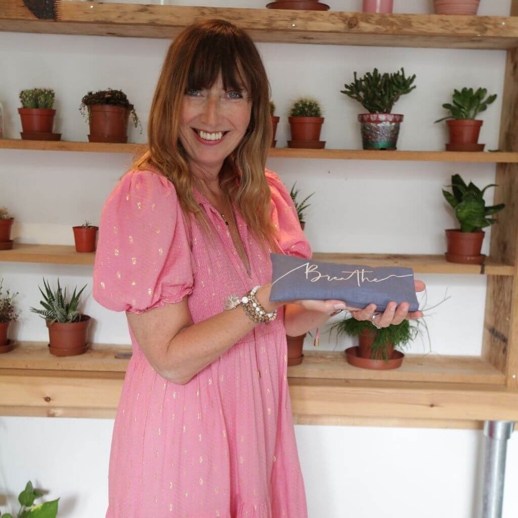 Lucy Edge in a pink dress holding a box with 'Breathe' embroidery in front of a wooden shelf with potted plants.