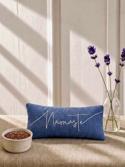 A blue rectangular eye pillow with 'Namaste' embroidered in white script, styled on a beige fabric surface with lavender in a glass vase and a small bowl of seeds against a sunlit cream wall.