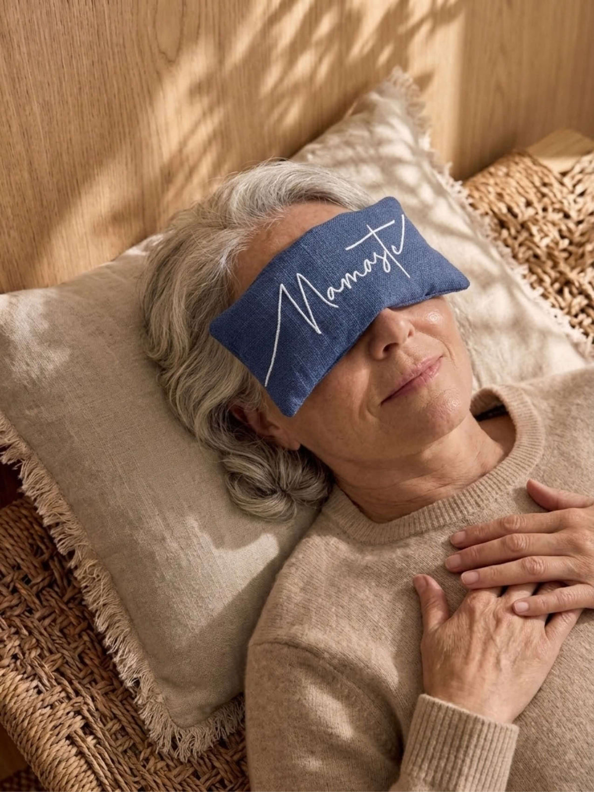 A person lying down on a woven chair with a beige pillow, wearing a blue eye pillow with the word 'Breathe' in white script.