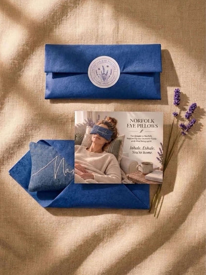 A styled flat-lay of a blue eye pillow set on a beige linen background with soft shadows. The set includes a blue envelope with a white circular seal, a blue eye pillow with white script embroidery, a printed insert card showing a woman wearing a blue eye mask, and dried lavender sprigs.