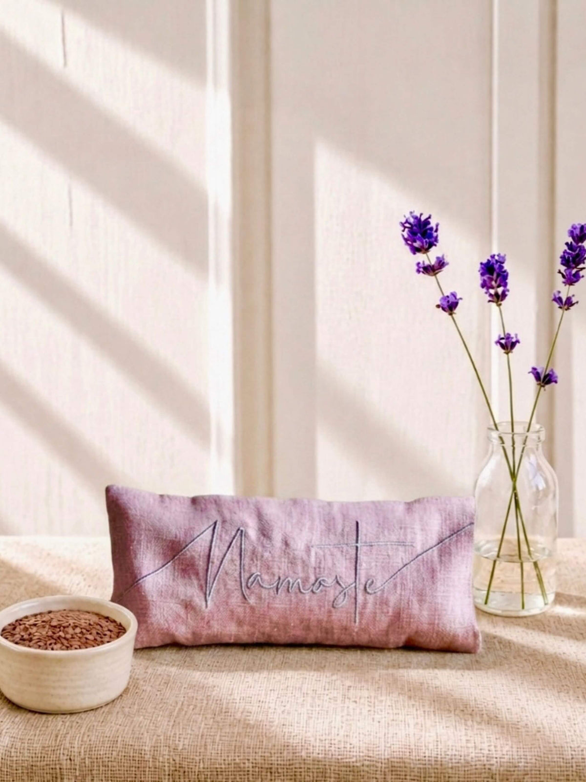 A rectangular pink eye pillow with 'Namaste' embroidered in grey script, styled on a beige linen surface with lavender stems in a glass vase and a small bowl of seeds against a sunlit cream wall.