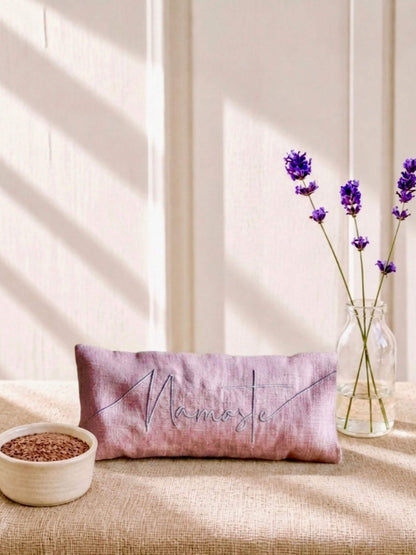 A rectangular pink eye pillow with 'Namaste' embroidered in grey script, styled on a beige linen surface with lavender stems in a glass vase and a small bowl of seeds against a sunlit cream wall.