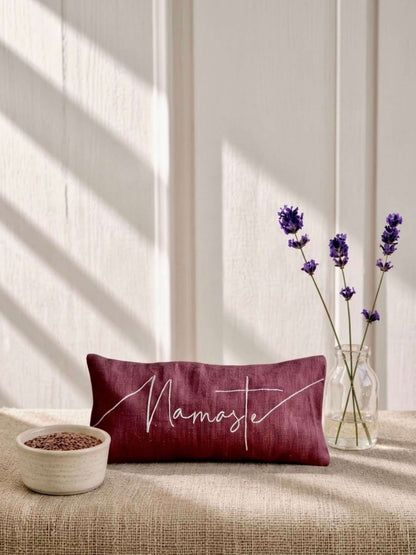 A rectangular burgundy eye pillow with 'Namaste' embroidered in white script, styled on a beige textured bench with lavender in a glass vase and a small bowl of seeds against a paneled wall with window shadow light.