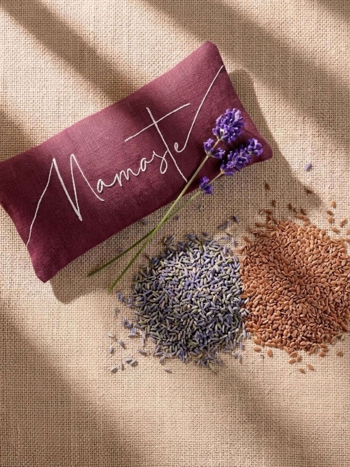 A rectangular lavender eye pillow in deep burgundy with 'Namaste' embroidered in white script, styled on a burlap surface with loose lavender buds and brown seeds under warm diagonal shadows.