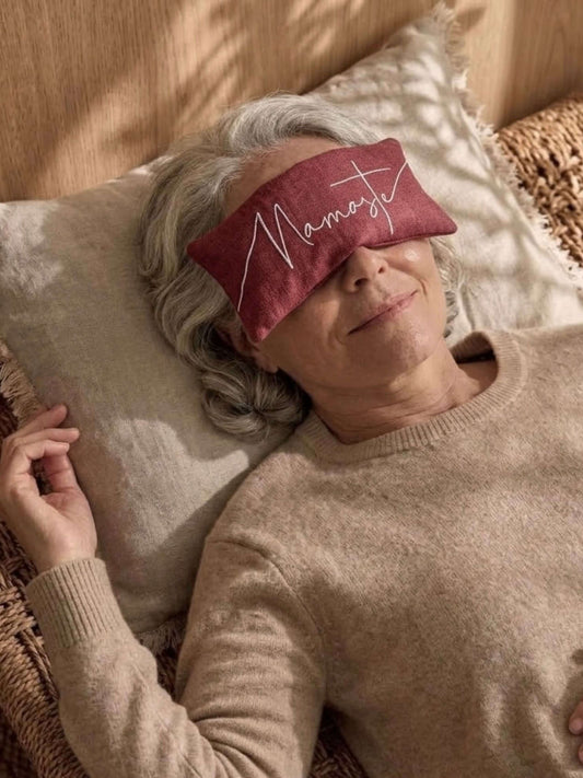 A woman lying down on a pillow wearing a red eye pillow with the word Namaste'' embroidered in white script.