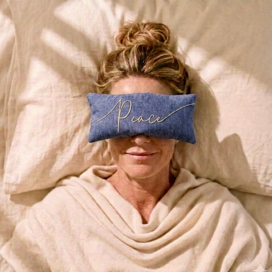 A woman lying in bed with a blue rectangular eye pillow featuring the word 'Peace' embroidered across the center. She is wrapped in a cream blanket, with pillows behind her and soft sunlight casting shadows across the scene.