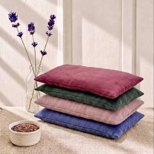 Four eye pillows in red, dark green, dusty pink, and blue are stacked on a beige fabric surface. A small white bowl with flaxseeds and a glass vase with purple lavender sprigs sit beside them against a cream panelled wall with soft window shadows.